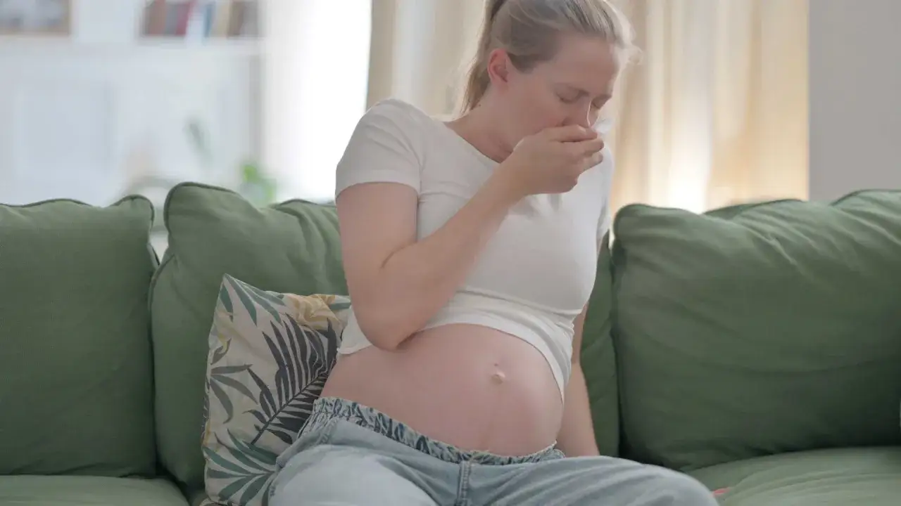Ciężarna kobieta siedzi na kanapie, zasłaniając usta chusteczką. Kichanie w ciąży to częsty problem.