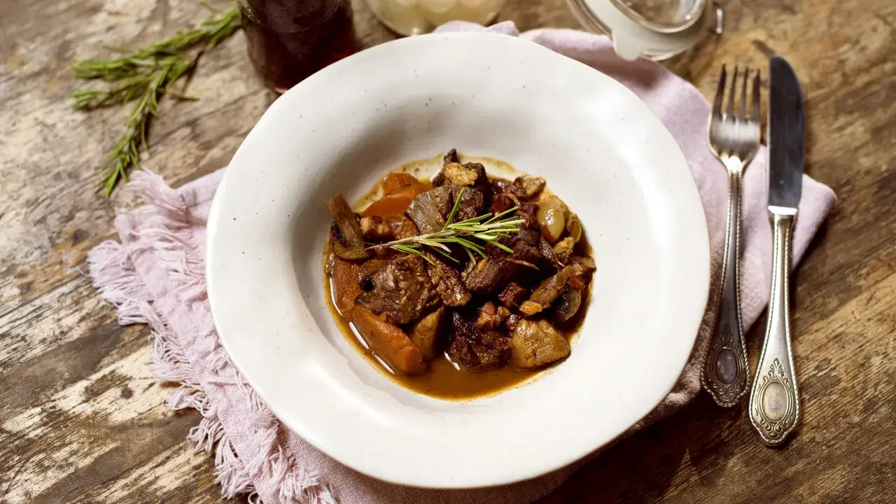 Aksamitny bœuf bourguignon z wołowiny, grzybów i marchewki, udekorowany gałązką rozmarynu, podany na białym talerzu.