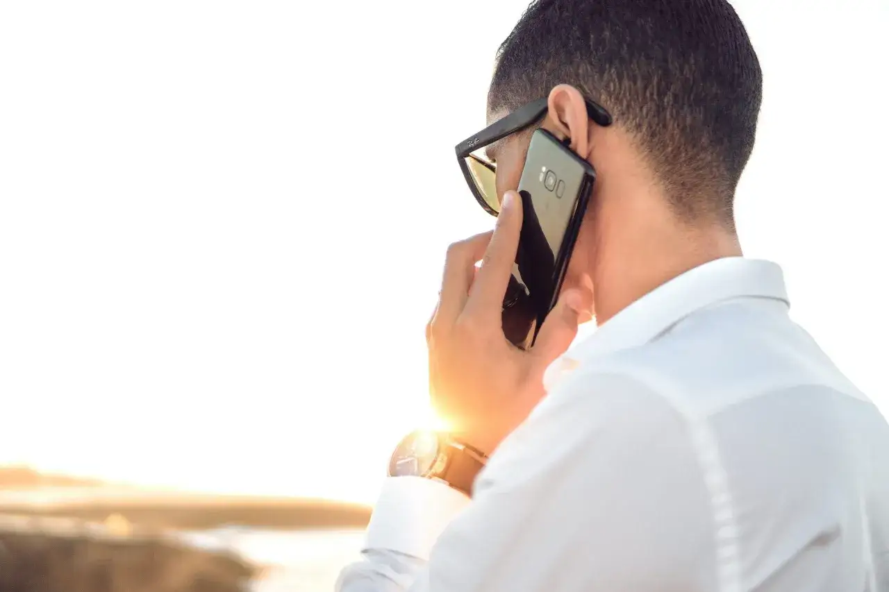 Mężczyzna w okularach przeciwsłonecznych i białej koszuli rozmawia przez telefon. Może to być cold caller, kt&oacute;ry pr&oacute;buje nawiązać kontakt.