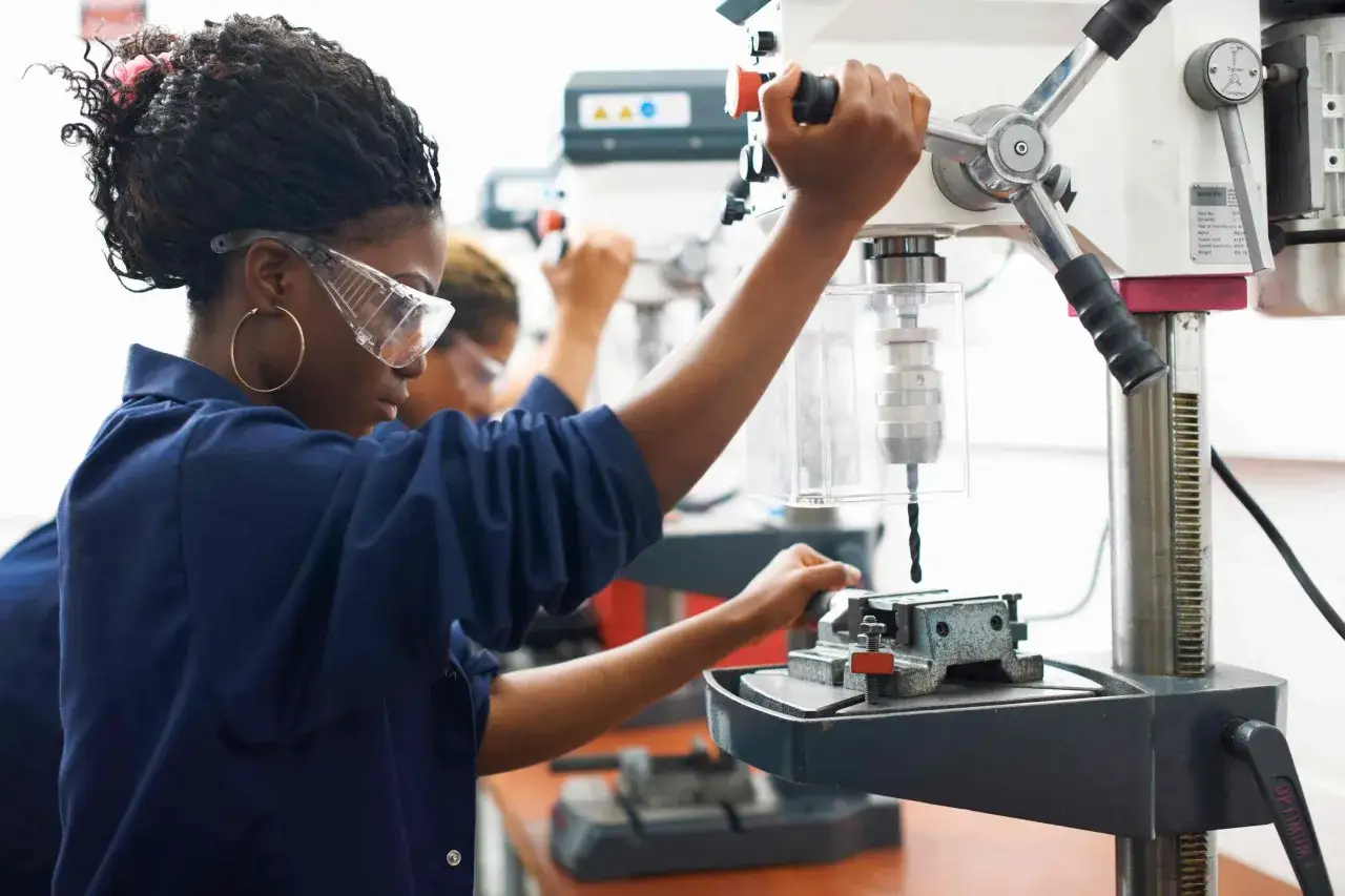 Junge Frau in Werkstatt mit Schutzbrille bedient Bohrmaschine. Einblicke in die berufsausbildung USA vergleichbar mit praktischer Lehre.
