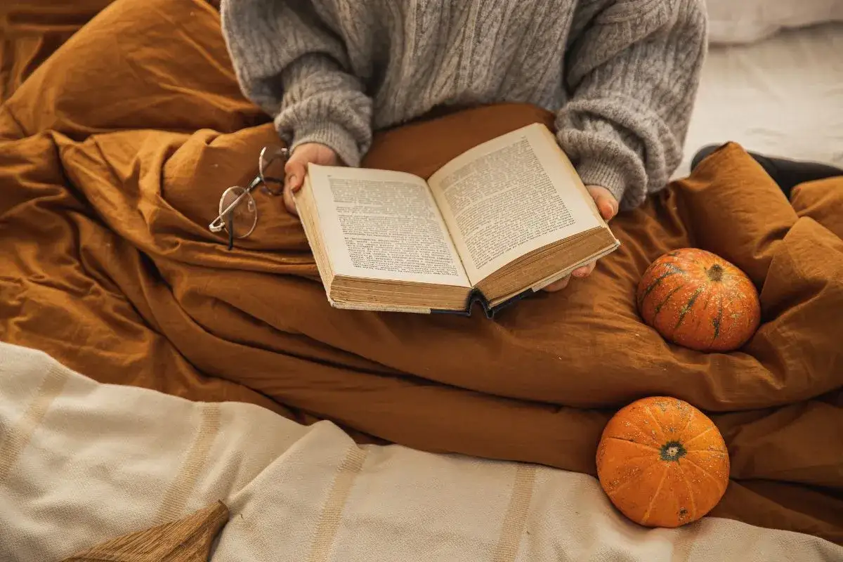 Osoba w swetrze czyta książkę, otoczona pomarańczowymi dyniami i kocami. Idealny nastrój na jesienne wieczory i straszne książki na Halloween.