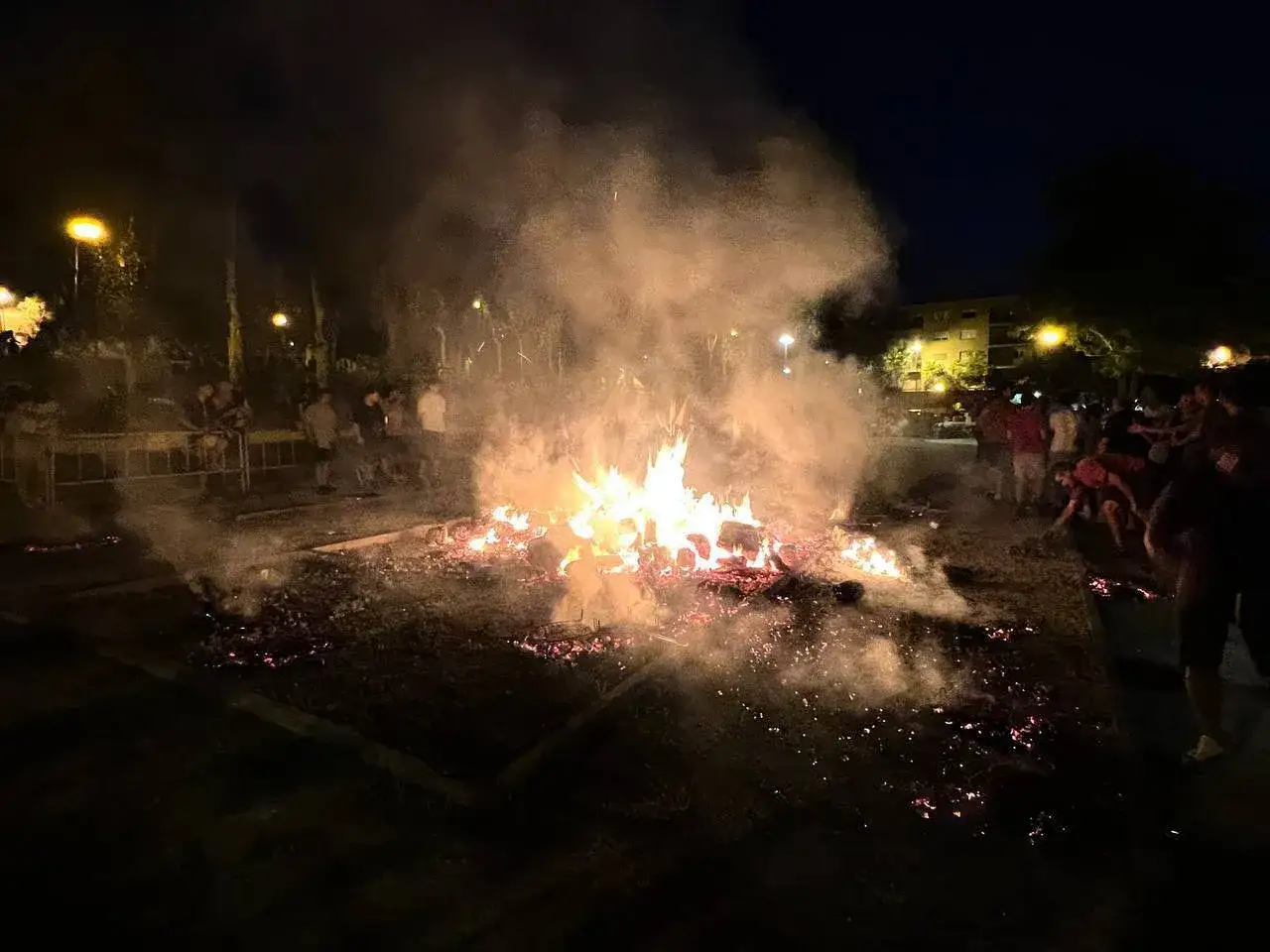 Noche de hogueras de San Juan en Zaragoza. Gente reunida alrededor de grandes hogueras ardiendo, con humo y chispas iluminando la oscuridad.