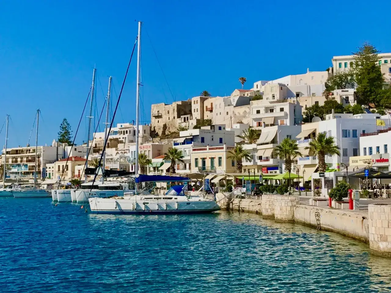 Segelbåtar i hamnen vid Naxos, Grekland, med vita hus och palmer längs kajen under en klarblå himmel.