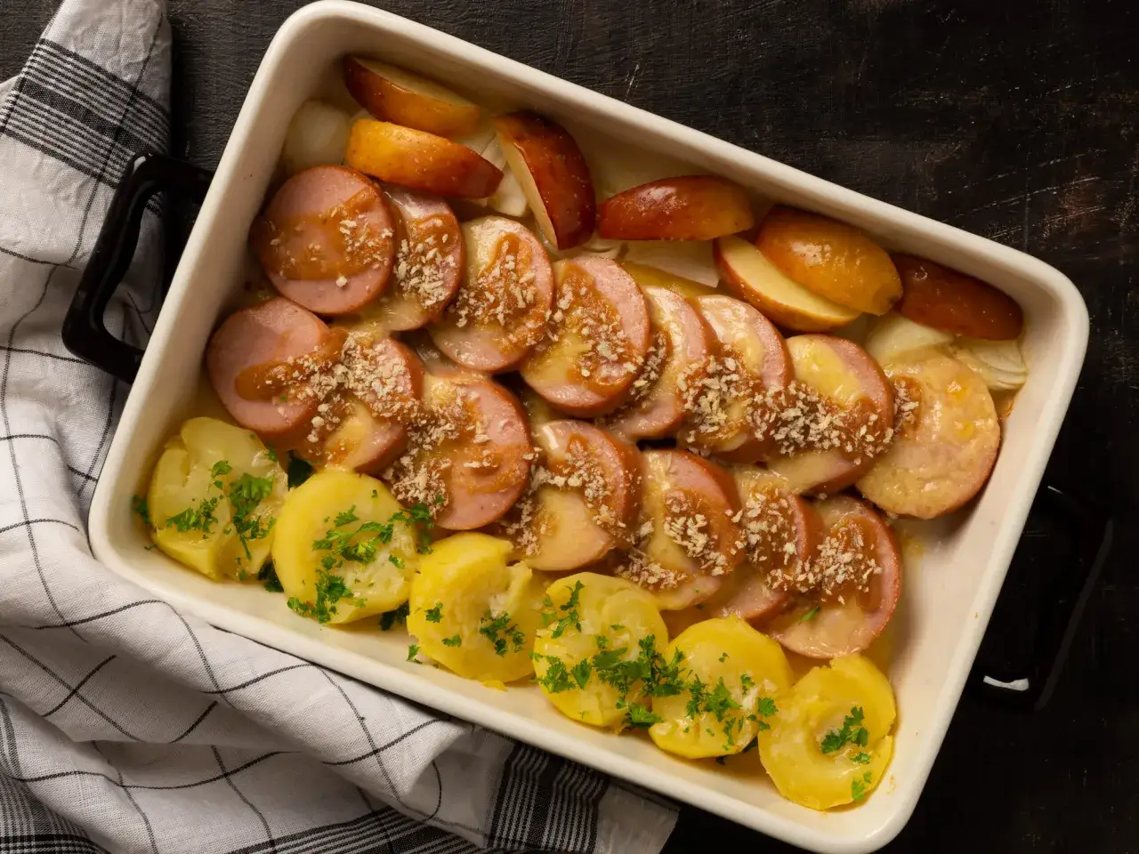 Ugnsbakad falukorv med potatis och äpple i en vit ugnsform. Garnerad med persilja och ströbröd.