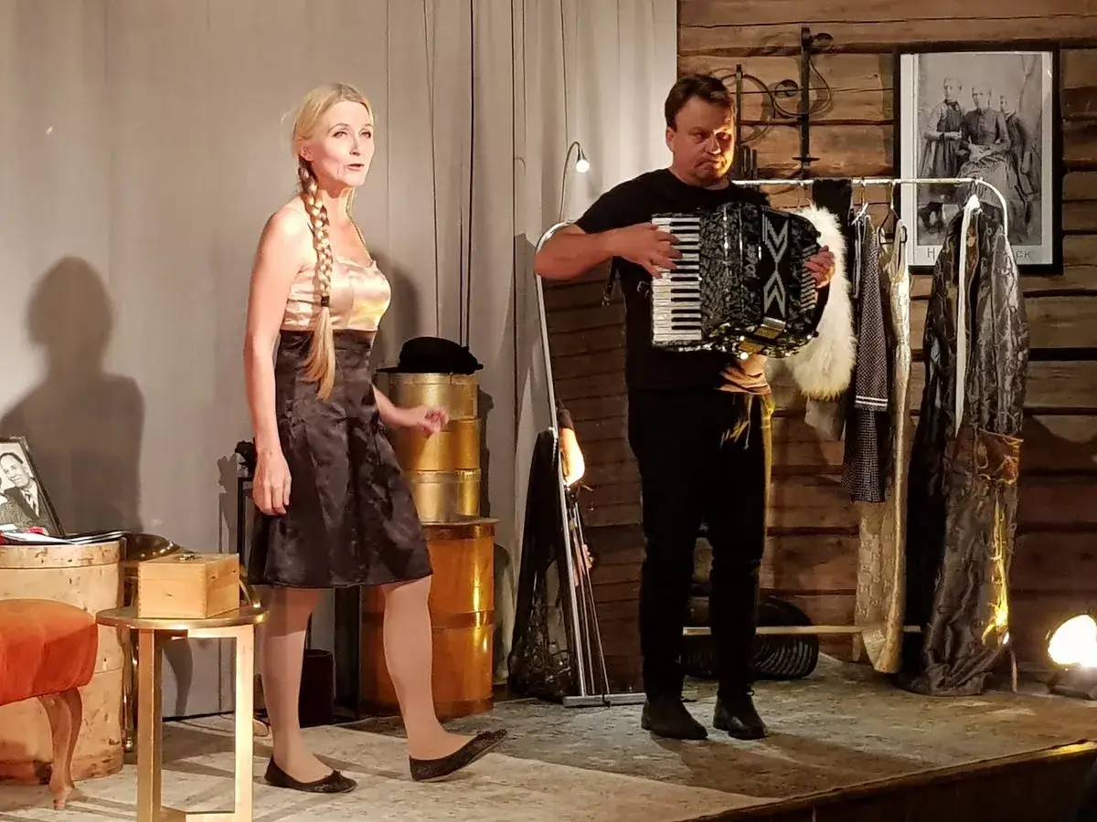En kvinna med flätat hår, klädd i en glänsande klänning, står på scenen. Bredvid henne spelar en man dragspel. Kanske en scen med svenska operasångerskor.