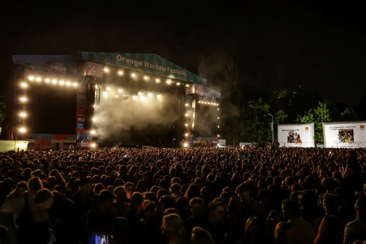 Tłum ludzi na Orange Warsaw Festival, jednym z największych festiwali w Polsce. Scena oświetlona, dym i światła tworzą atmosferę.