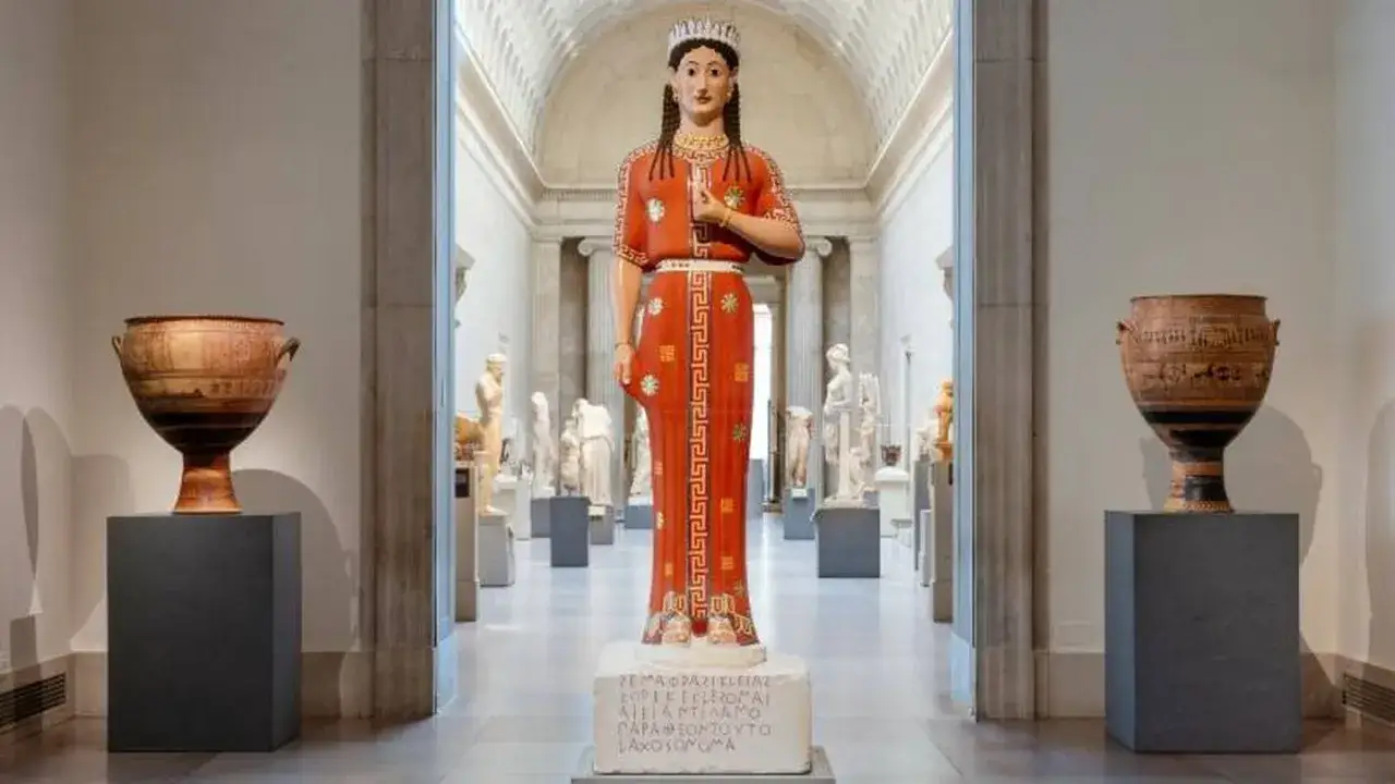 Una colorida escultura griega de una mujer con un vestido rojo, flanqueada por vasijas antiguas en un museo.
