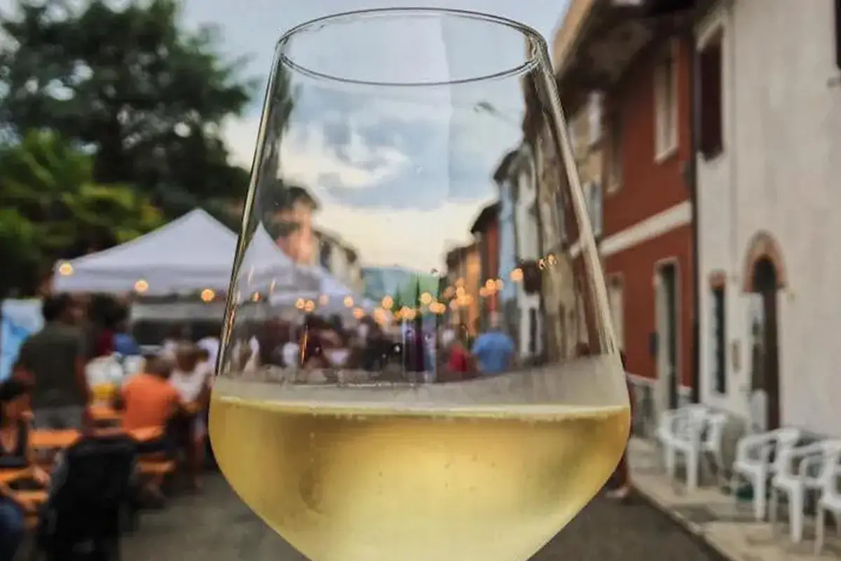Un bicchiere di vino bianco riflette una festa di paese nel Val Tidone, con luci soffuse e gente che passeggia.