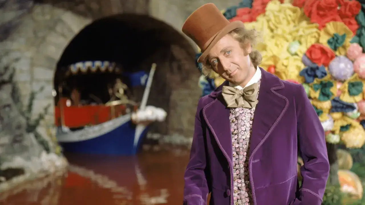 Gene Wilder, i sin ikoniska lila sammetsrock och hatt, st&aring;r vid en chokladflod med en b&aring;t.