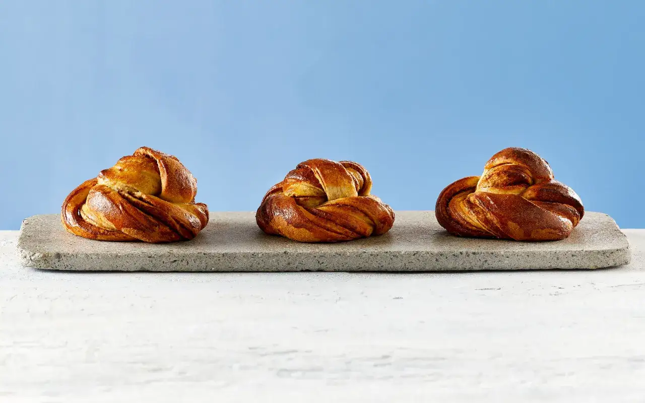 Tre gyllene laktosfria kanelbullar ligger p&aring; en stenplatta mot en bl&aring; bakgrund.