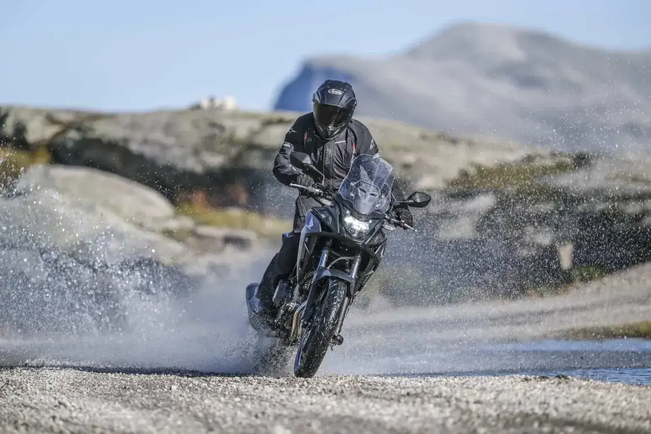 Motocyklista w kasku i kurtce jedzie motocyklem Honda CB500X przez wodę, tworząc rozpryski.