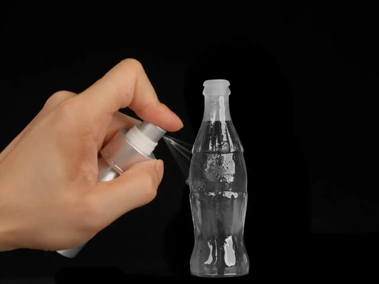 A hand sprays a clear 3d print of a Coca-Cola bottle with a fine mist.
