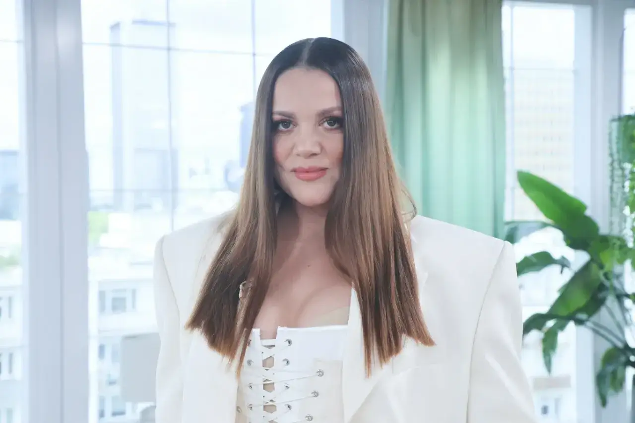 Dominika Gawęda w białej marynarce i gorsetowym topie, z długimi, brązowymi włosami. Wygląda jakby właśnie skończyła karmić dziecko.
