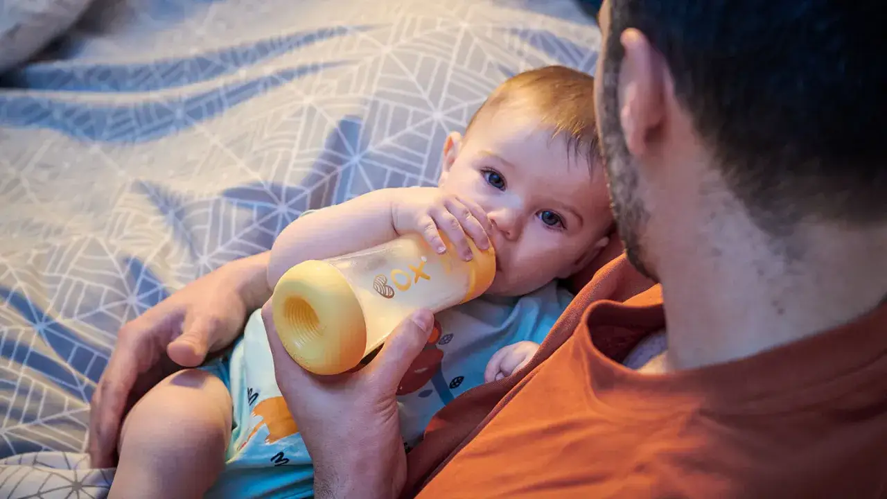 Un padre tiene un biberón per dare da mangiare al suo neonato, un momento tenero su un letto con lenzuola a fantasia.