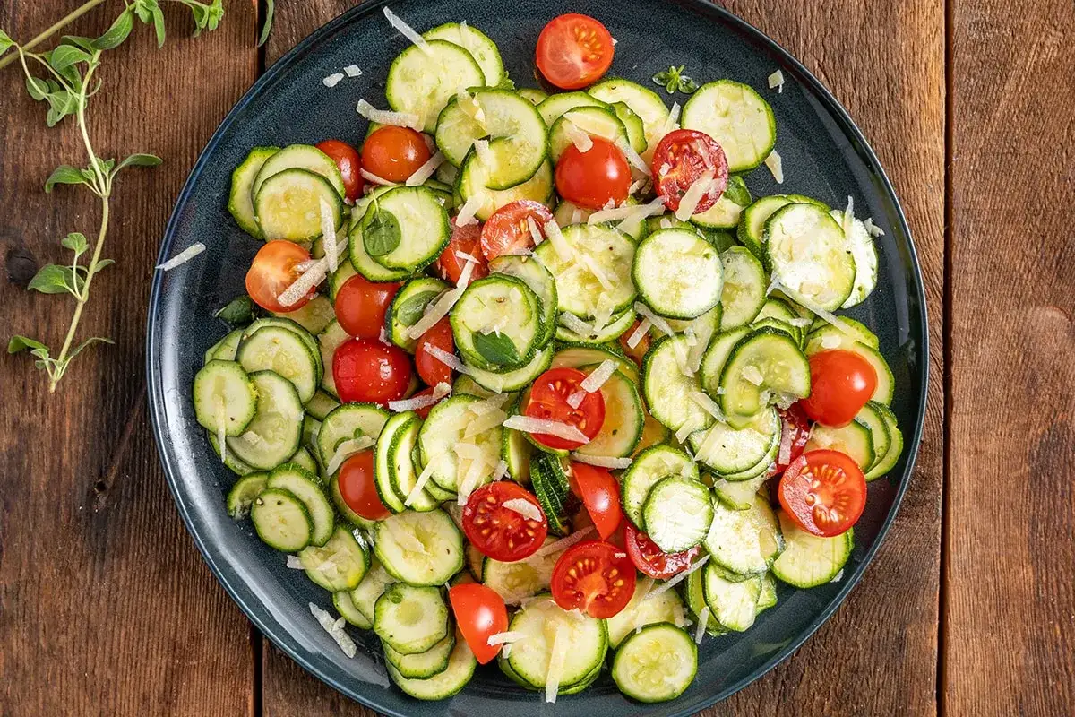 Deliziosa insalata di zucchine fresche, pomodorini ciliegino e scaglie di parmigiano, guarnita con erbe aromatiche.