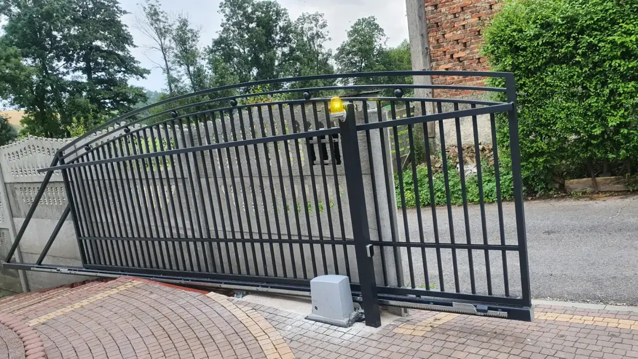 Czarna, metalowa brama wjazdowa z żółtym sygnalizatorem świetlnym, częściowo otwarta. Obok niej widoczny napęd.