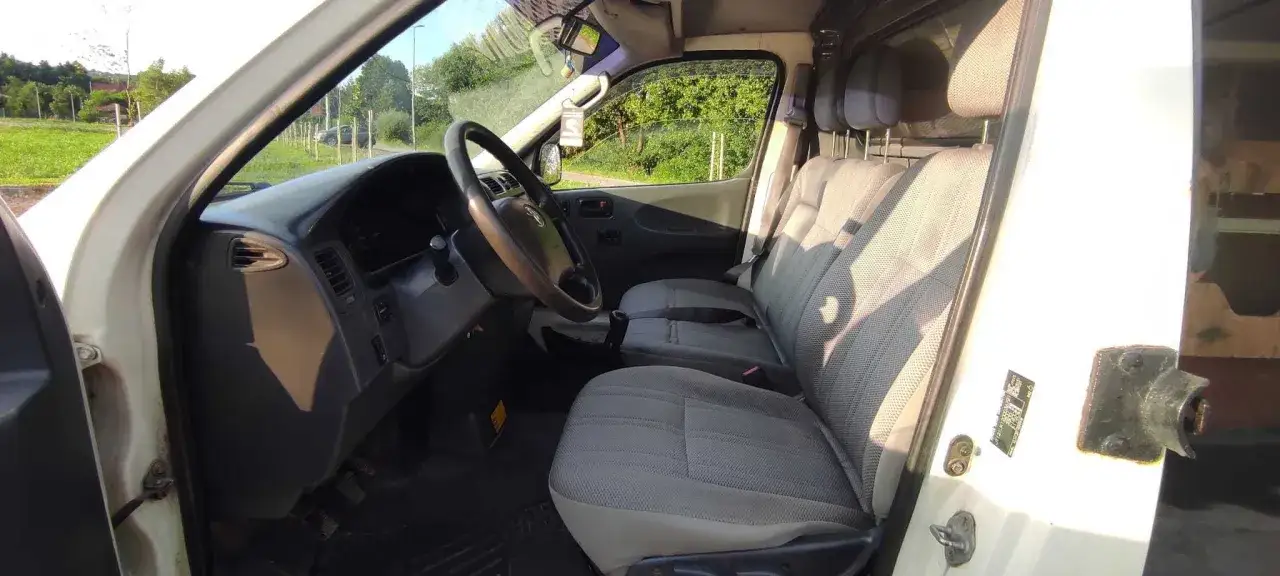Wnętrze białej Toyoty Hiace z szarymi siedzeniami i kierownicą. Widok z boku, przez otwarte drzwi.