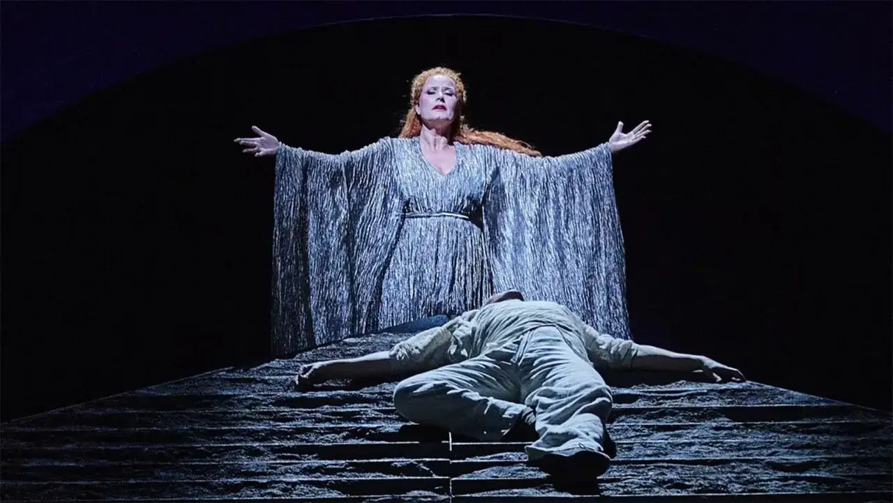 Kobieta w srebrnej sukni z rozłożonymi ramionami góruje nad leżącym u jej stóp mężczyzną. Dramatyczna scena operowa, pełna emocji, jakby zapowiadała potężną arię.