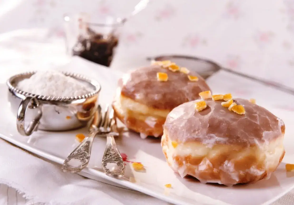 Stary przepis na pączki, dwa puszyste przysmaki z lukrem i kandyzowaną skórką pomarańczową, obok cukru pudru i dżemu.