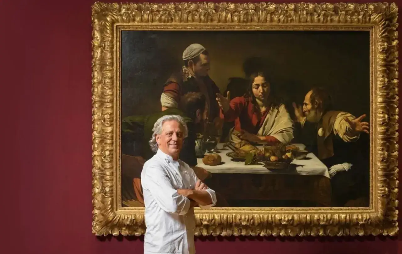 Giorgio Locatelli, chef stellato Michelin, posa davanti a un dipinto storico di un banchetto.