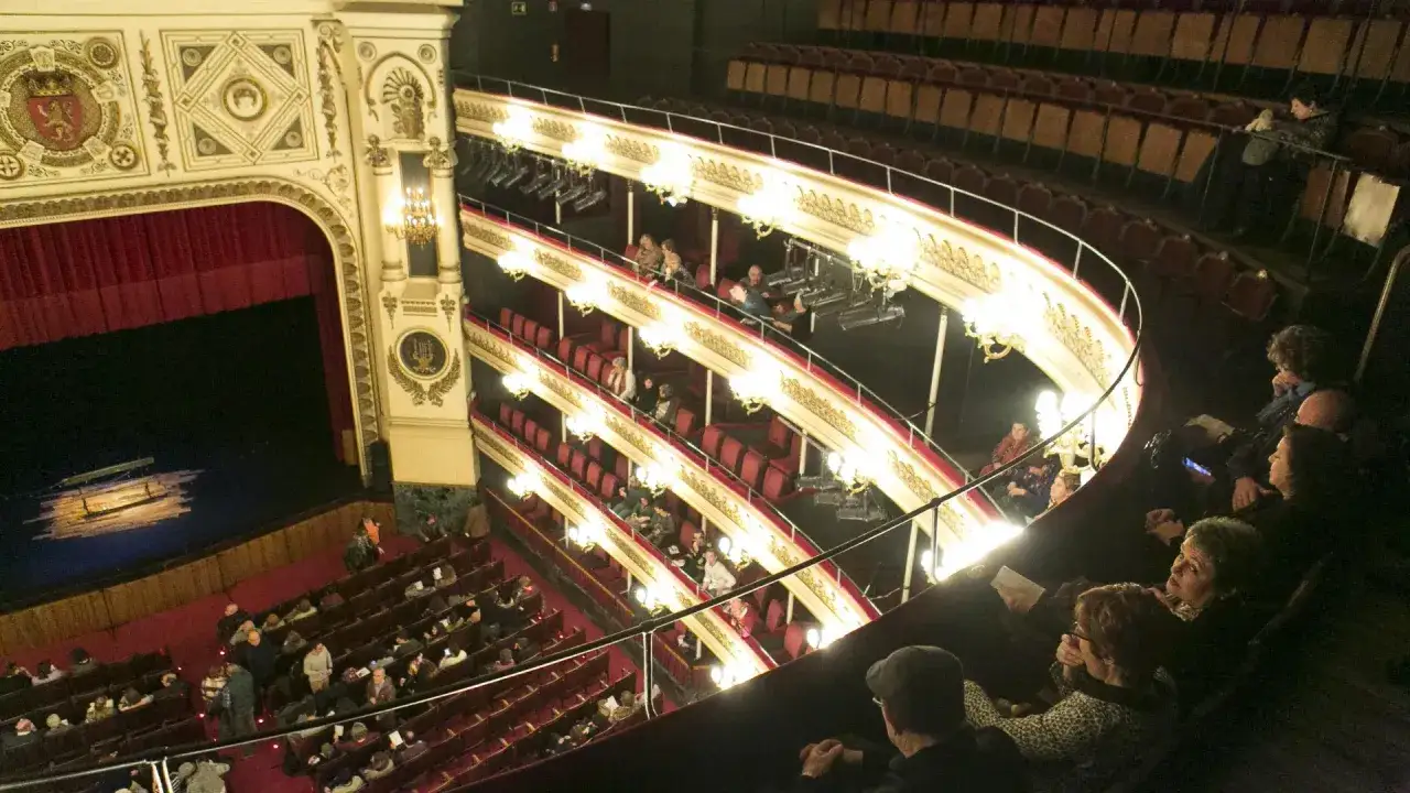 Interior de un teatro con público esperando el inicio de musicales en Zaragoza. Butacas rojas y doradas, escenario con telón y luces.