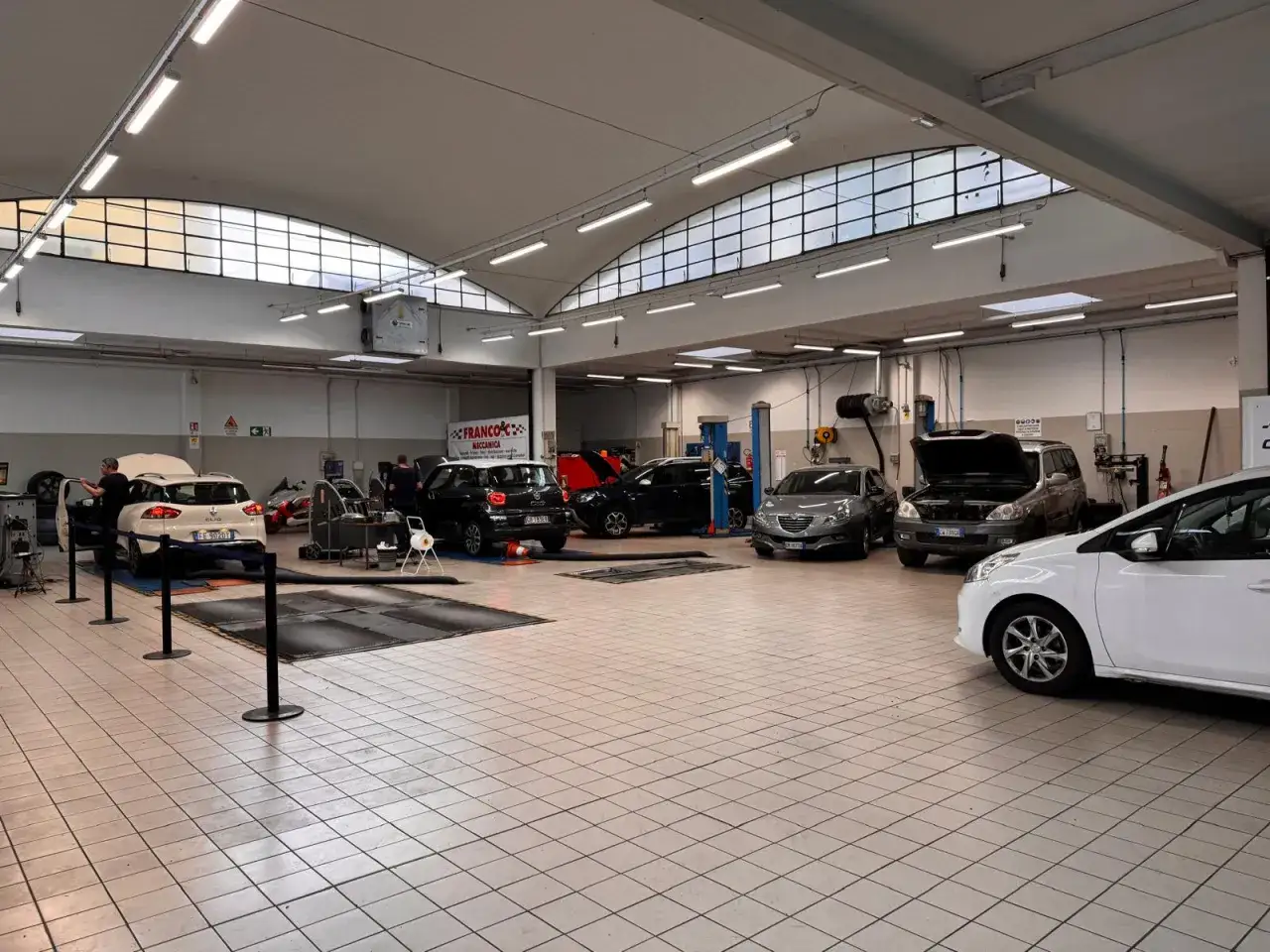 Officina meccanica con auto in revisione. Un meccanico lavora su una microcar bianca, mentre altre auto attendono il loro turno.