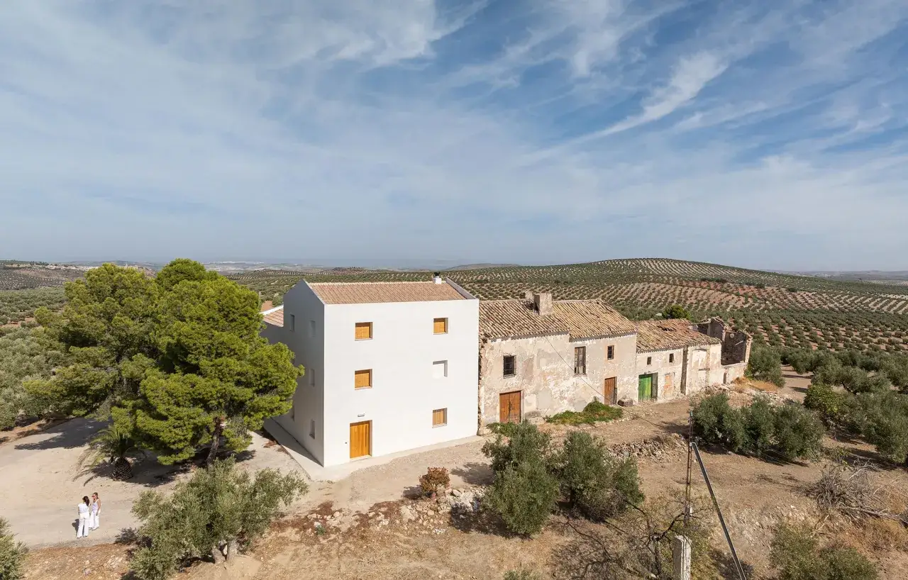 El cortijo Bacares, con su nueva edificaci&oacute;n blanca junto a las ruinas, se alza entre olivares bajo un cielo azul con nubes.
