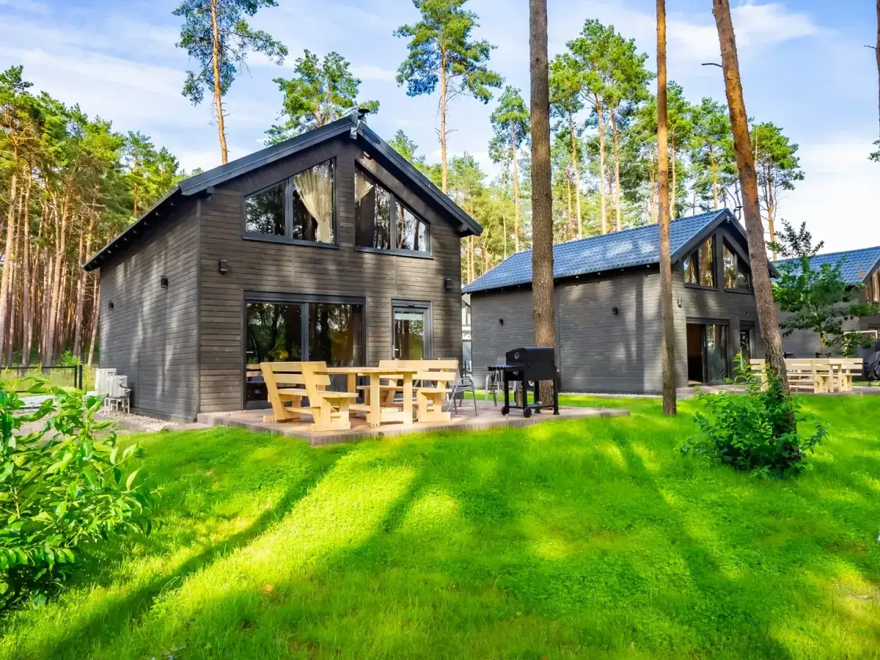 Nowoczesne lubiatów domki nad jeziorem w otoczeniu sosnowego lasu. Drewniane meble i grill czekają na gości.