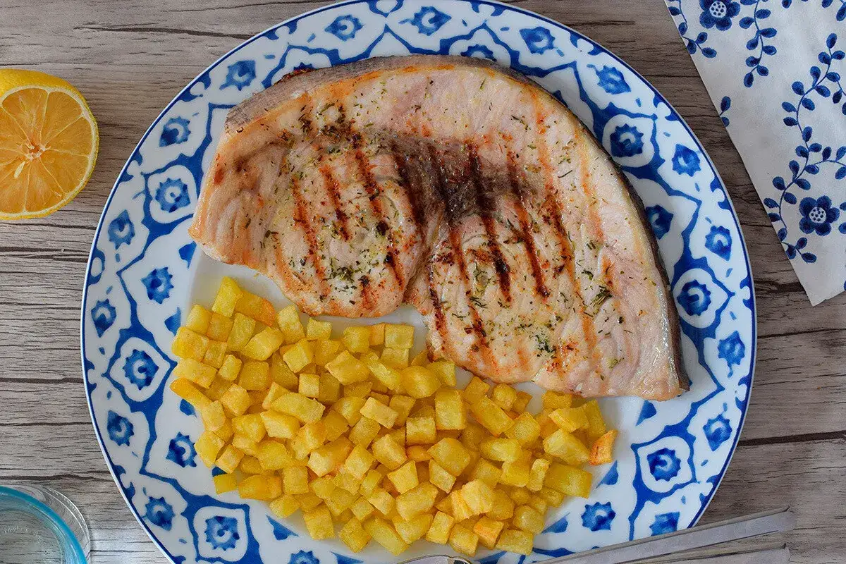 Delicioso emperador a la plancha con patatas fritas crujientes, servido en un plato con dise&ntilde;o azul y blanco. Un toque de lim&oacute;n completa esta apetitosa presentaci&oacute;n.