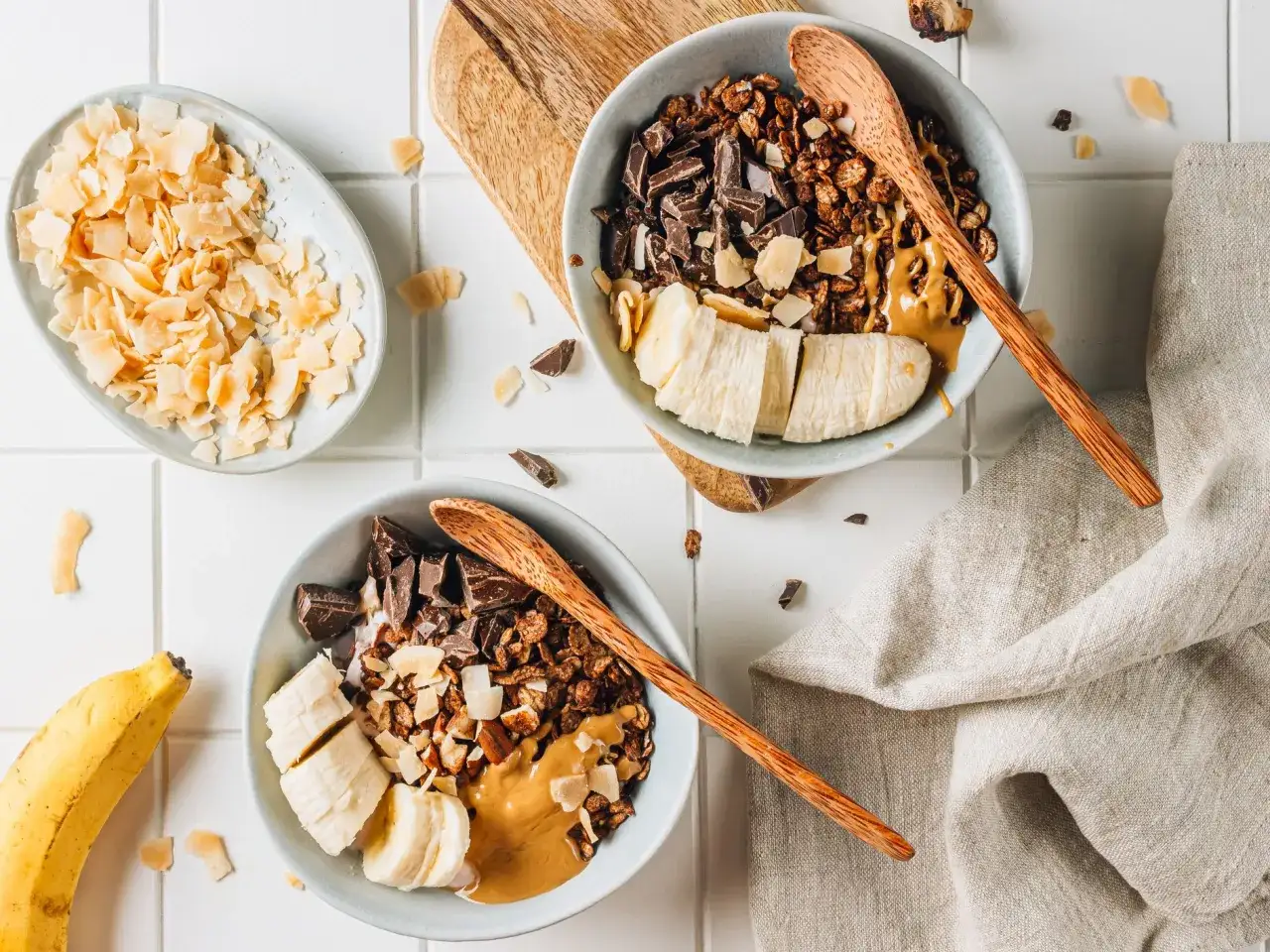 Due ciotole con granola fatta in casa, banana a fette, cioccolato fondente e burro d'arachidi. Cocco a scaglie a lato.