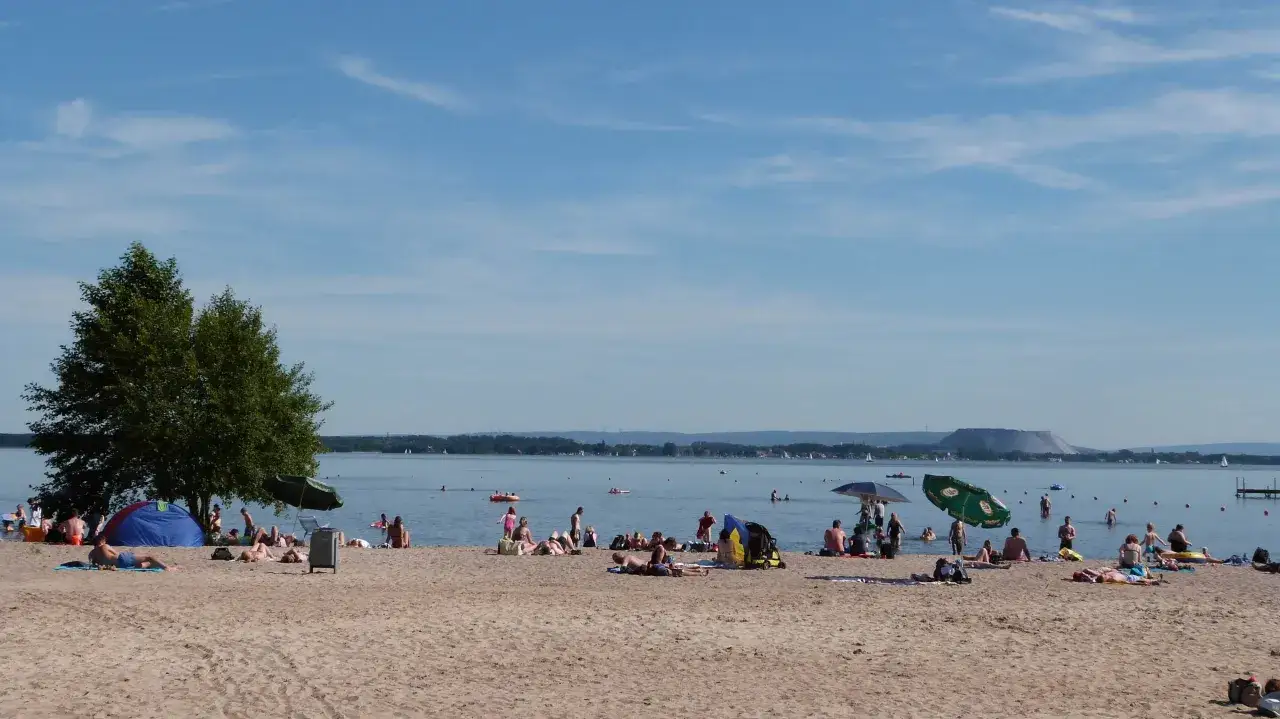 Menschen entspannen sich am Sandstrand des Steinhuder Meeres. Boote segeln auf dem Wasser, w&auml;hrend im Hintergrund eine sanfte H&uuml;gellandschaft zu sehen ist.