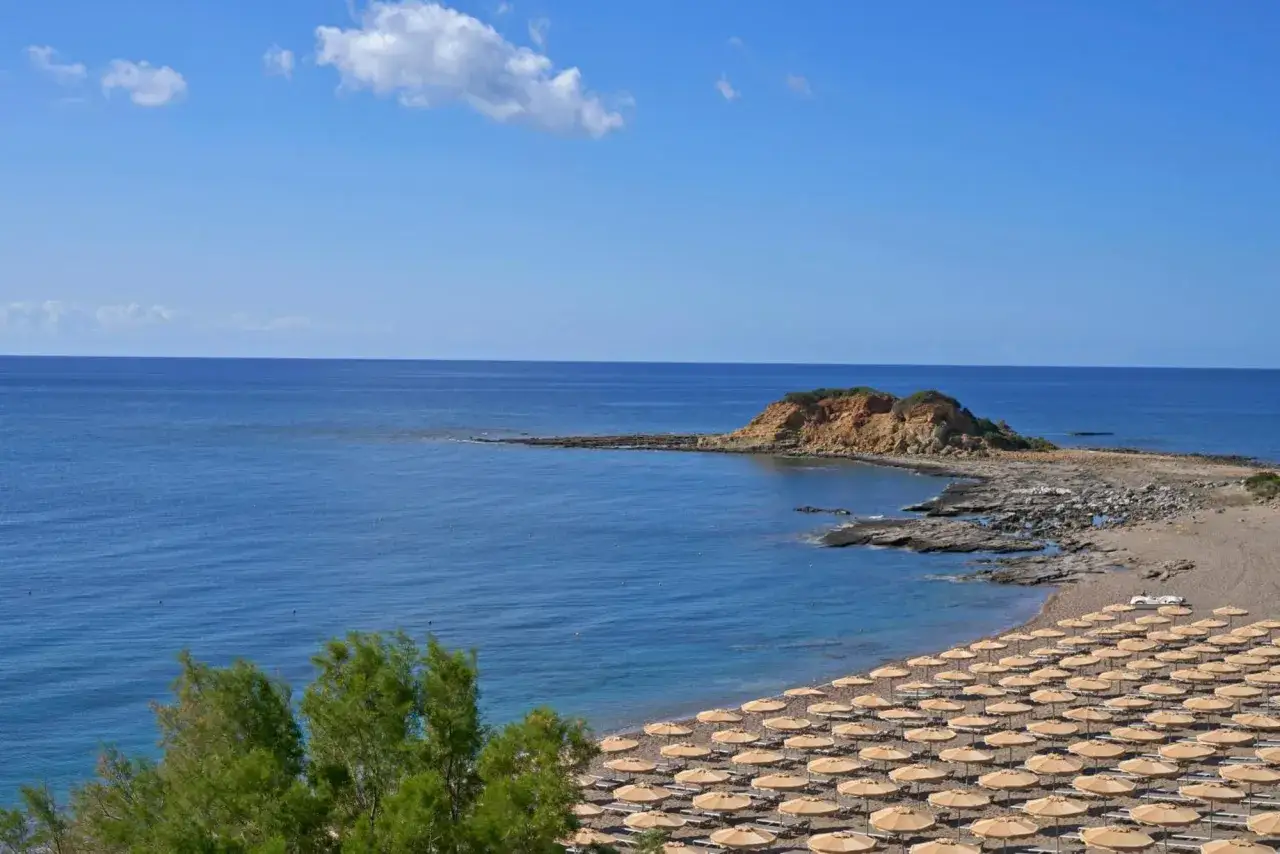 Strand mit vielen Sonnenschirmen und Liegen am Rodos Princess Beach Hotel. Das Meer ist blau, im Hintergrund eine felsige Insel.