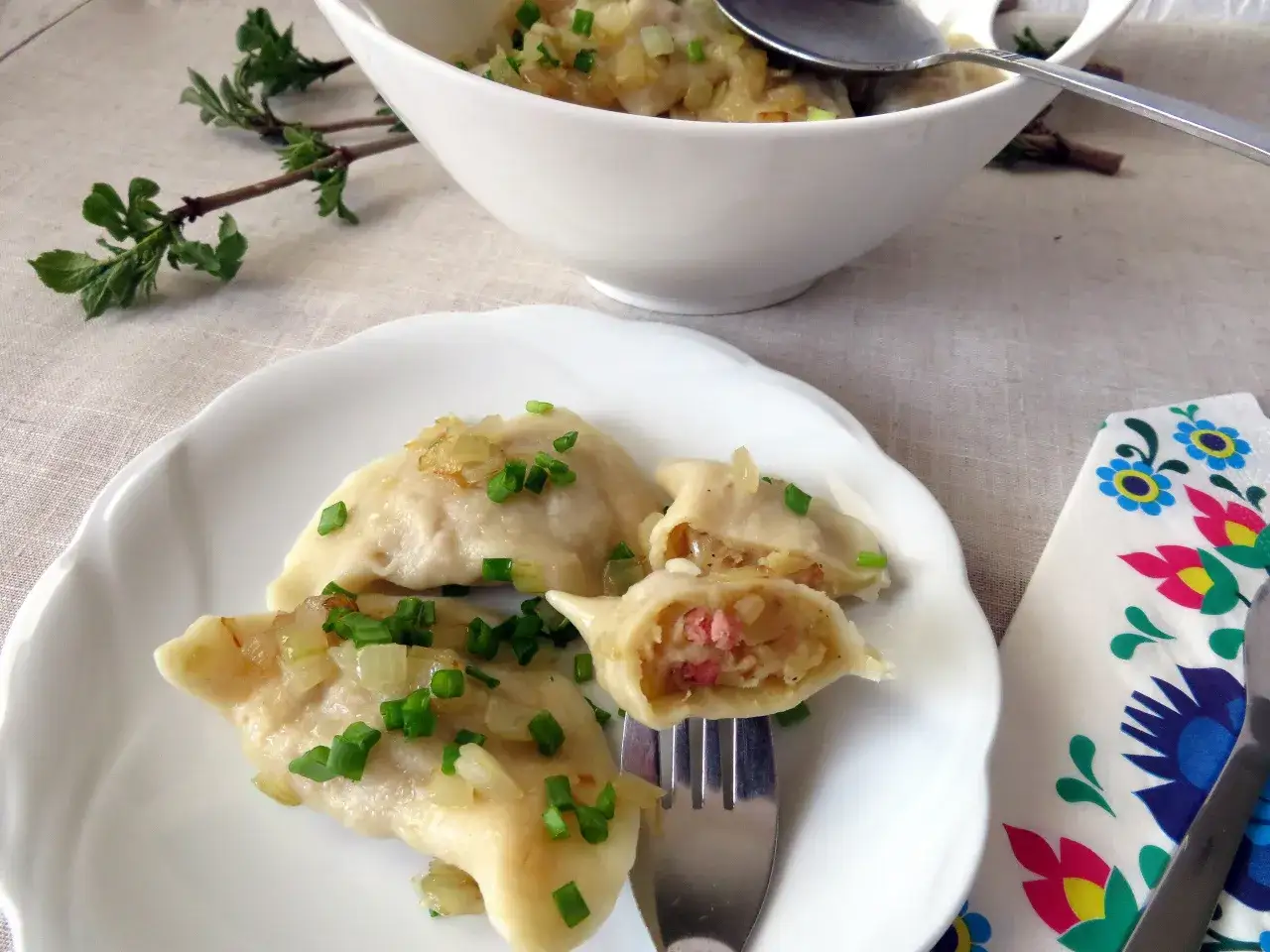 Pyszne pierogi z ziemniakami i boczkiem, posypane szczypiorkiem i cebulką, na białym talerzu.