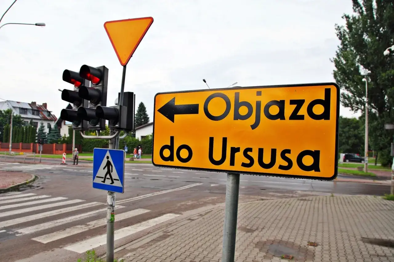 Pomarańczowy znak drogowy z czarną strzałką w lewo informuje o objazd do Ursusa. Obok sygnalizacja świetlna i znak przejścia dla pieszych.