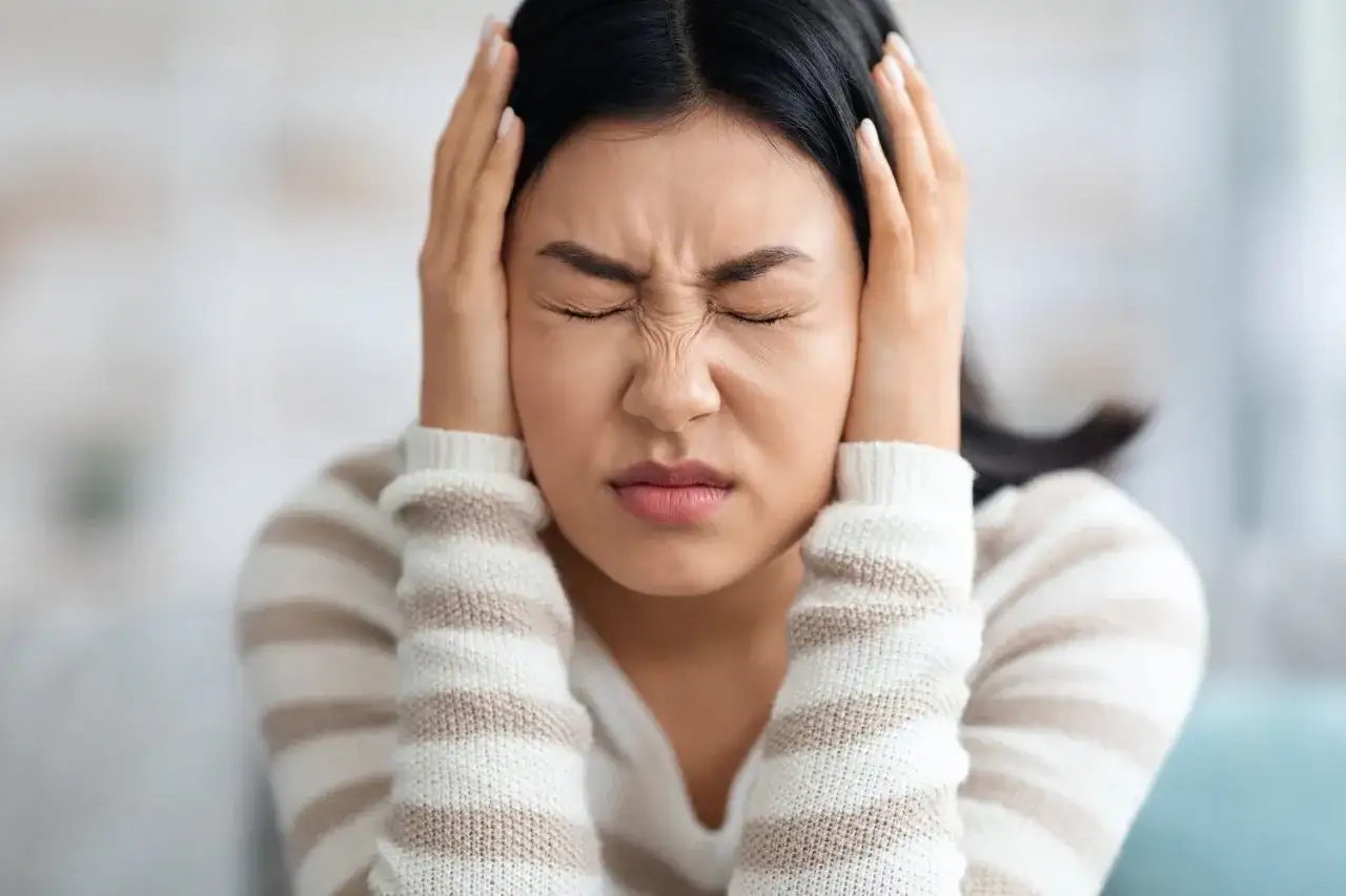 Frau mit geschlossenen Augen und Händen an den Ohren, die unter Tinnitus leidet. Osteopathie kann helfen.