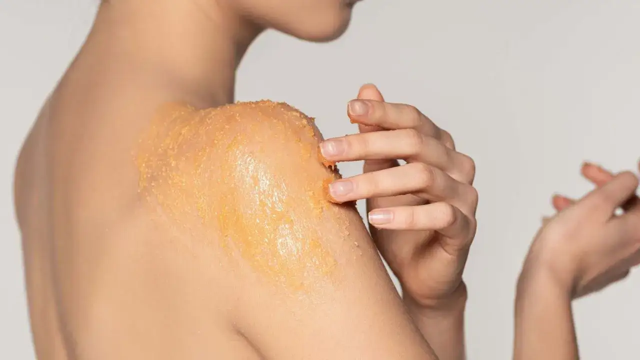 Mani che applicano scrub corpo sulla spalla, un gesto di cura per una pelle liscia e luminosa.