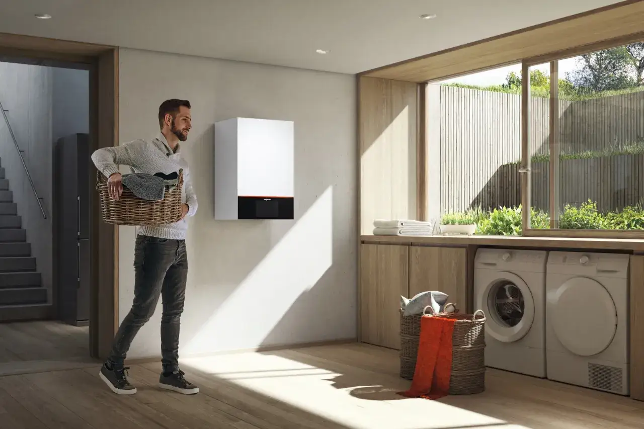Hombre con cesta de ropa junto a una de las calderas m&aacute;s peque&ntilde;as del mercado, en un cuarto de lavander&iacute;a moderno.