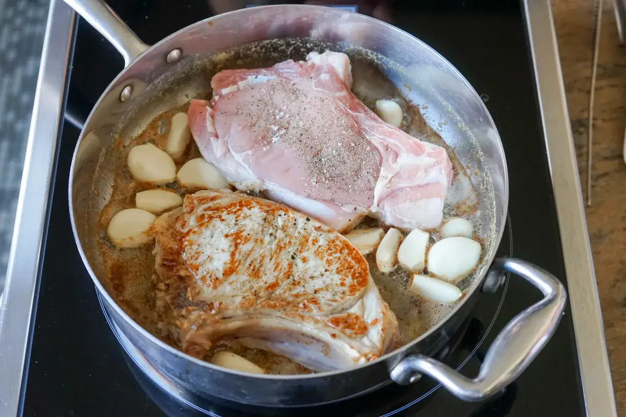 Deux côtes de veau crues et une dorée cuisent dans une poêle avec de l'ail et du beurre.