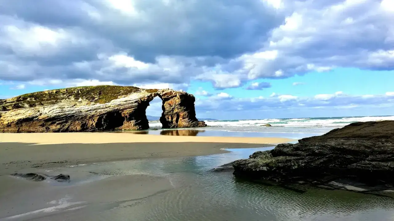 Arco natural en la playa de Las Catedrales, norte de Espa&ntilde;a en verano. Arena dorada, mar agitado y cielo nublado.