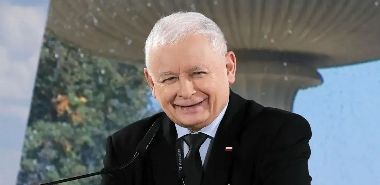 Jarosław Kaczyński w garniturze, uśmiechnięty, na tle zieleni i nieba.