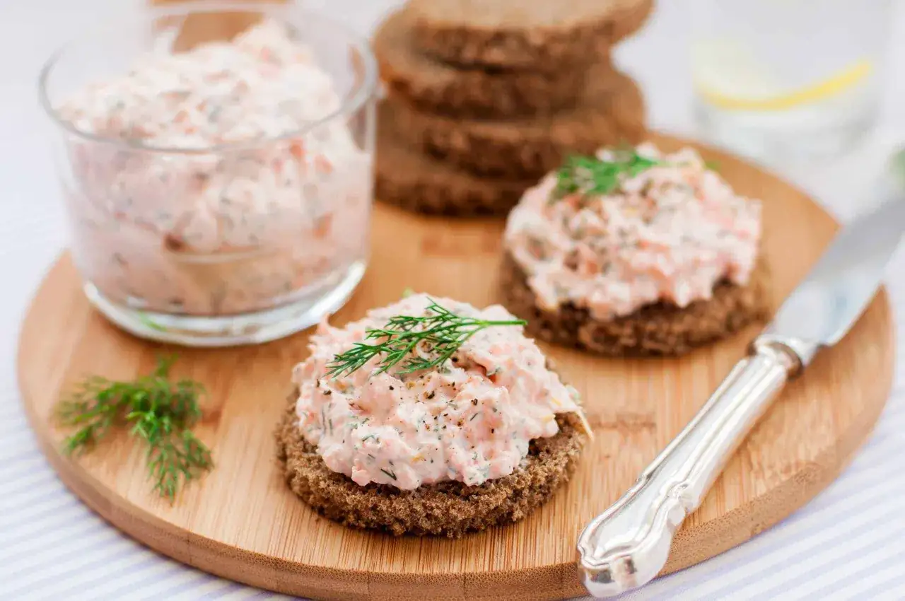 Deux toasts de pain noir garnis de rillettes de saumon, décorés d'aneth. Un verre rempli de rillettes est visible à gauche.