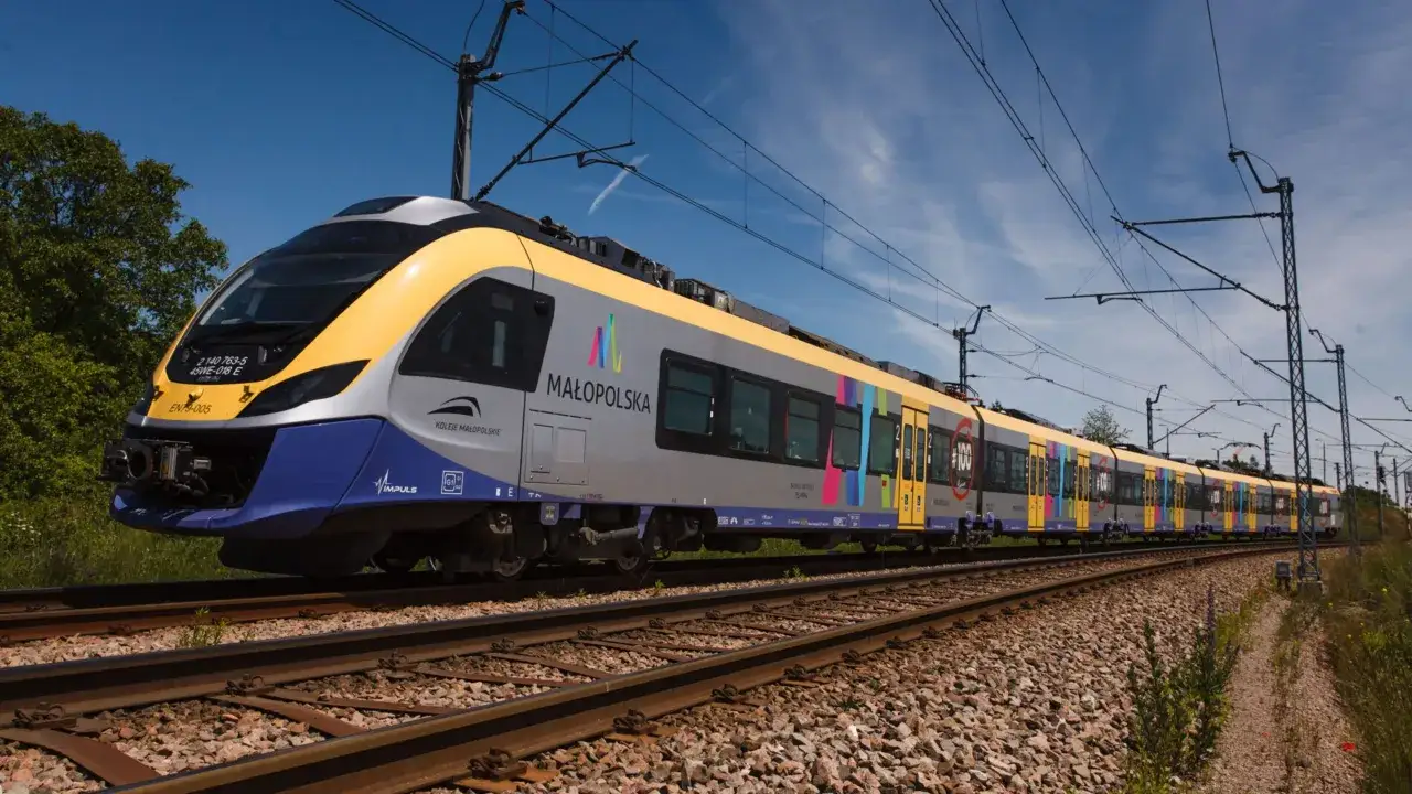 Nowoczesny pociąg 45WE-018 E Kolei Małopolskich z kolorowymi pasami jedzie po torach w słoneczny dzień.