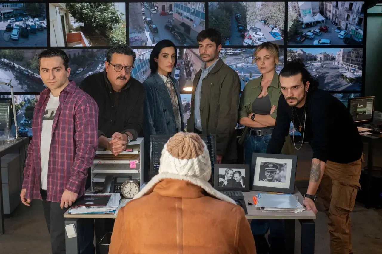 Il cast di 100 years in un centro di controllo, con schermi che mostrano immagini di sorveglianza.