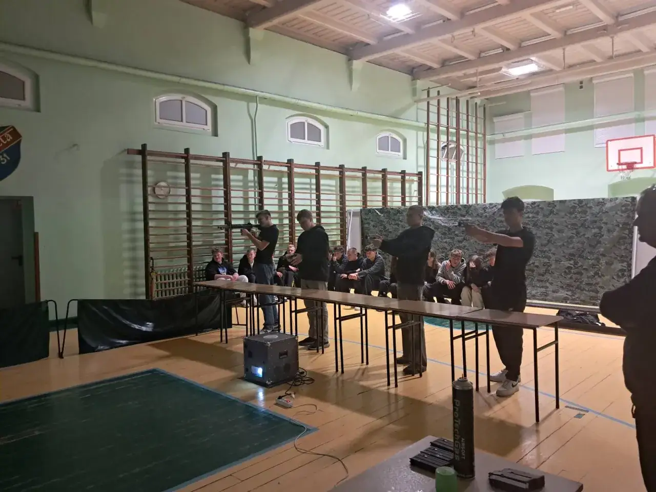 Uczniowie szkoły branżowej 2 stopnia ćwiczą strzelanie z broni pneumatycznej na sali gimnastycznej.
