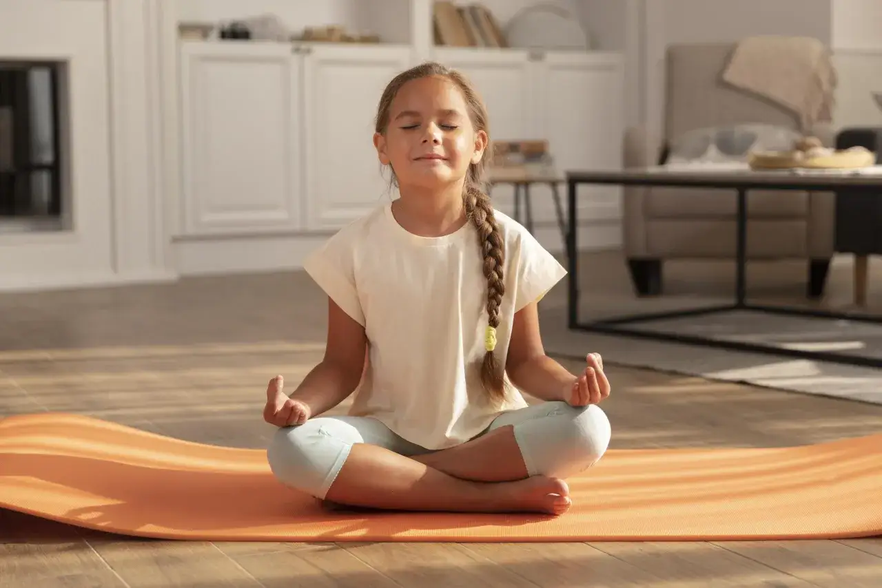Niña practica posturas yoga niños en una esterilla naranja, con los ojos cerrados y las manos en mudra.