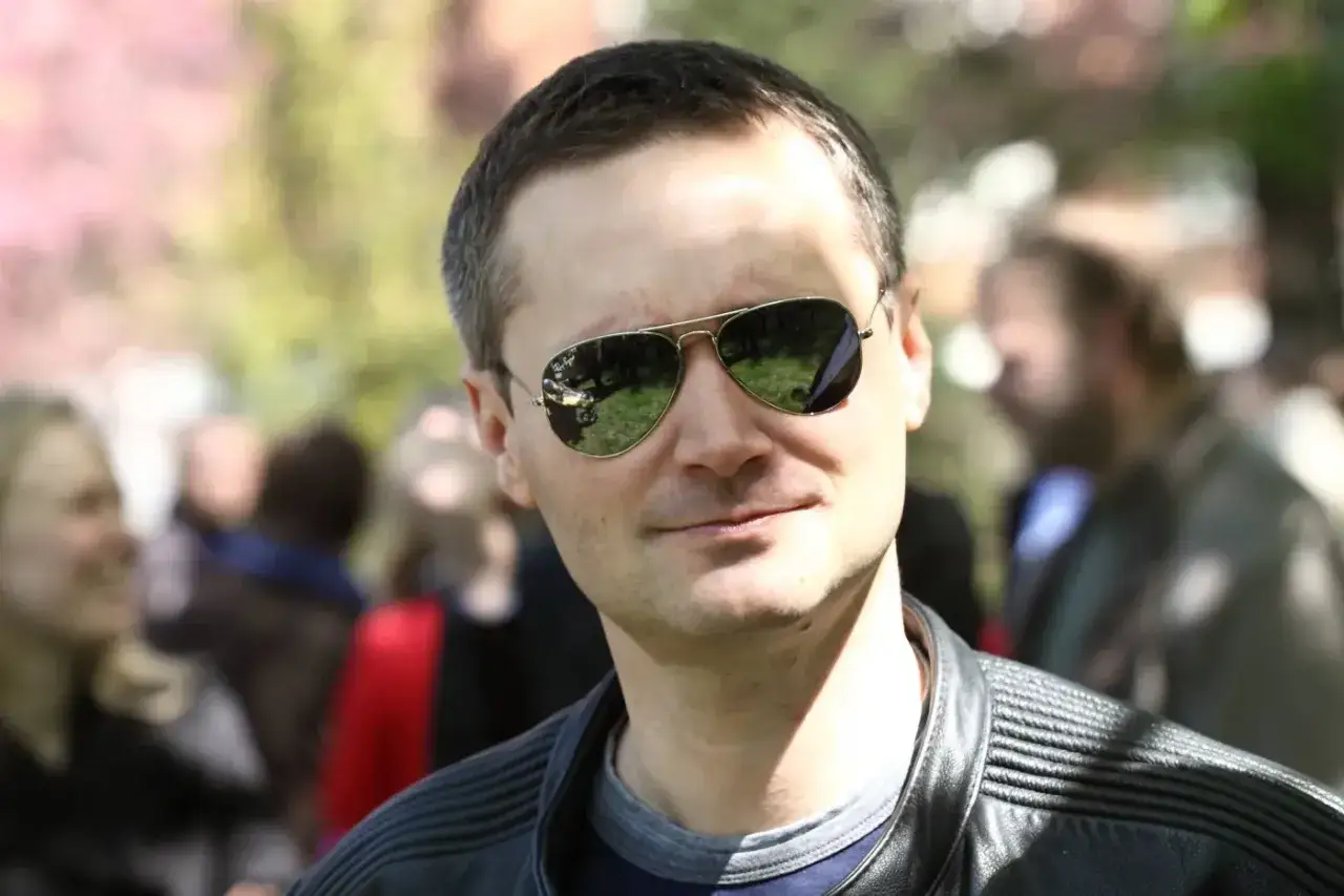 Jarosław Wałęsa w okularach przeciwsłonecznych, w tle rozmyte postacie. Jego wiek nie jest widoczny, ale wygląda na dojrzałego mężczyznę.
