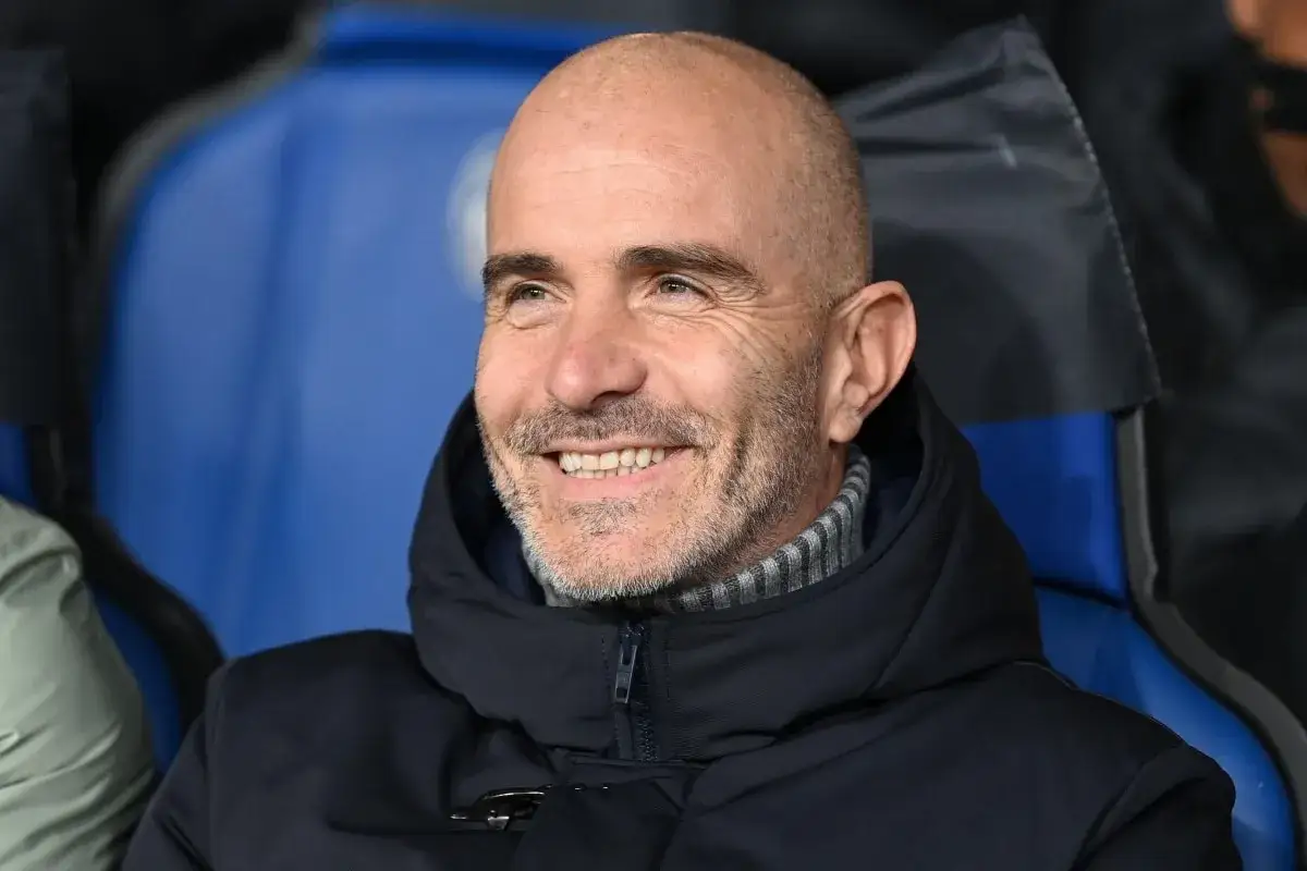 Uśmiechnięty trener Manchester City, z siwą brodą i łysą głową, siedzi na ławce w ciemnej kurtce.