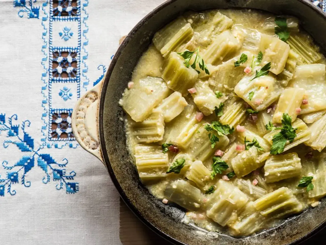 Cardo en salsa cremosa con trocitos de jamón y perejil, servido en un cuenco sobre un mantel bordado. Un plato reconfortante con el toque crujiente de las almendras.