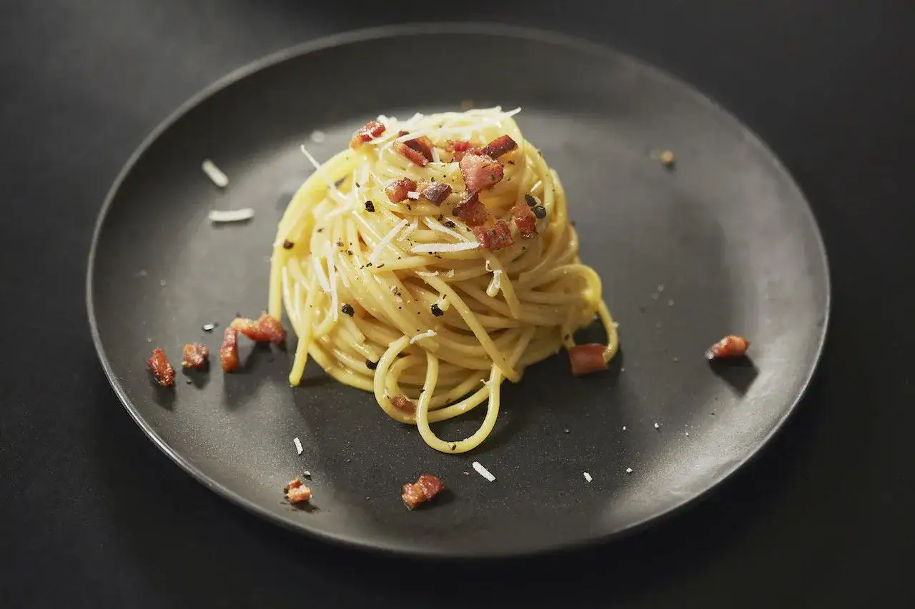 Spaghetti carbonara sur une assiette noire, parsemés de lardons croustillants et de copeaux de parmesan. Une recette simple et délicieuse.