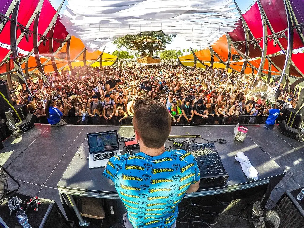 DJ na scenie, tłum szaleje przy muzyce EDM. Kolorowe konstrukcje nad głowami tworzą festiwalową atmosferę.
